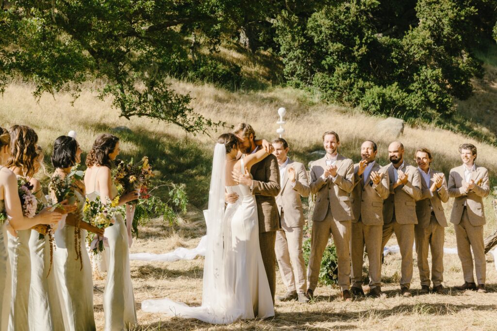 men's beige color suits. outdoor wedding ceremony. The photo shows a man and woman getting married with groomsmen wearing beige suits and bridesmaids wearing light green dresses.
