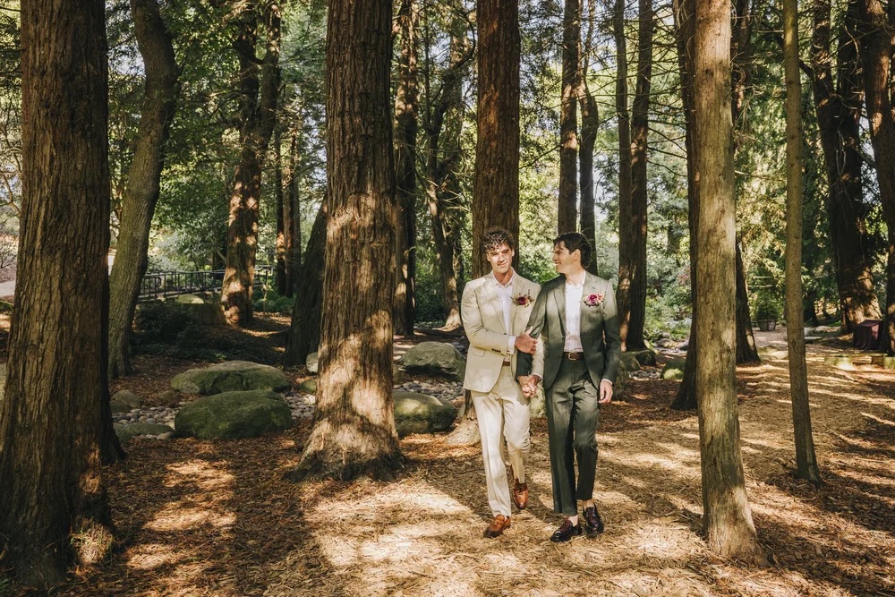 two grooms holding hands and walking through the woods. one is wearing a beige suit and the other is wearing a green suit.